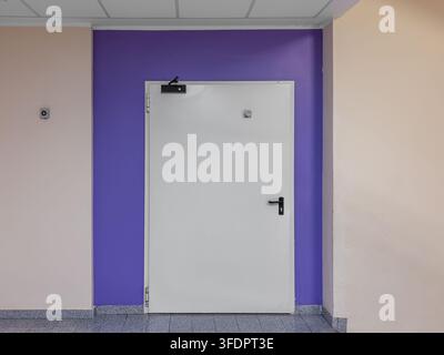 Single leaf white metal fire exit door with black handle and automatic closer hydraulic system installed on a purple painted wall section in a modern Stock Photo