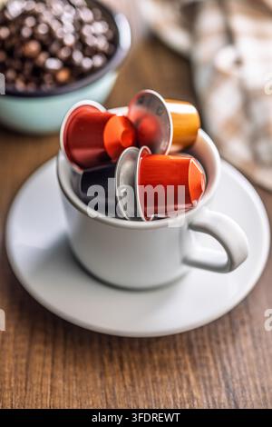 Bright metallic coffee capsules arranged inside a white cup and saucer on a wooden table, with a blurred bowl of coffee beans and warm linens conveyin Stock Photo