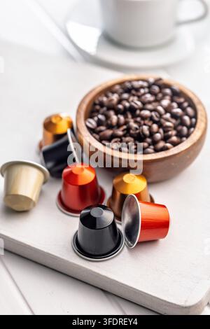 Colorful single‑serve coffee capsules scattered beside a wooden bowl filled with roasted coffee beans and a blurred white cup, suggesting a morning ri Stock Photo