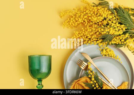 Spring table setting with mimosa flowers ceramic plates golden cutlery yellow napkin and green glass on pastel background bright festive composition Stock Photo