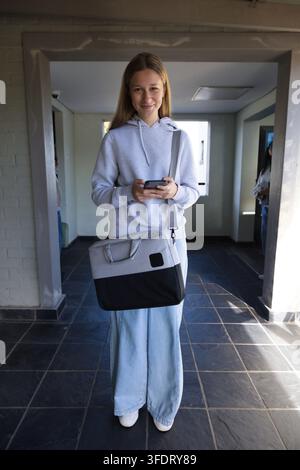 Female youth standing in tiled entry holding smartphone low, wearing grey hoodie and crossbody bag Stock Photo