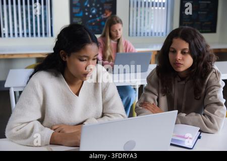 Diverse female students collaborating on silver laptop at classroom desks with notebooks and pen Stock Photo