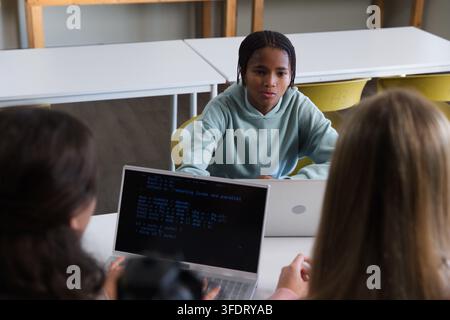 Diverse classmates studying and discussing code on laptops with terminal at school by white tables Stock Photo