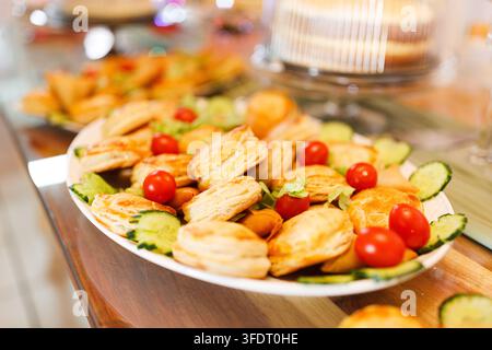 Close-up of assorted golden-brown savory puff pastries arranged on a serving plate with fresh lettuce, cucumber slices and cherry tomatoes on a wooden Stock Photo