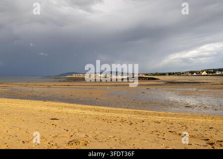 Penrhyn Bay North Wales beach at low tide with broad sandy shoreline shallow water distant coastal homes hills and dark dramatic clouds stretching acr Stock Photo