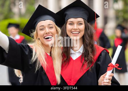 Two cheerful young ladies girlfriends in graduation clothes taking selfie, happy female students showing diplomas and smiling at camera, closeup portr Stock Photo