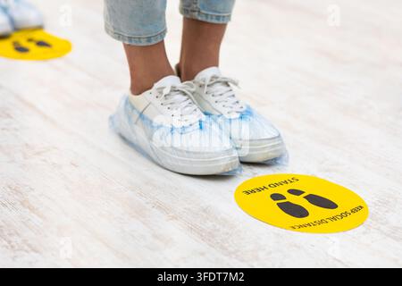 Keep Safe Distance. Teen Girl's Feet With Shoes Covers Standing Near Social Distancing Warning Sign On Floor In Queue Line Indoors. Covid-19, Coronavi Stock Photo