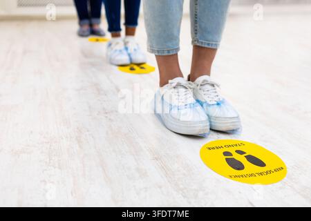 Social Distancing. People Standing In Queue, Closeup Of Feet In Shoes Covers And Warning Signs On Floor Indoors. Covid-19, Coronavirus Prevention Back Stock Photo