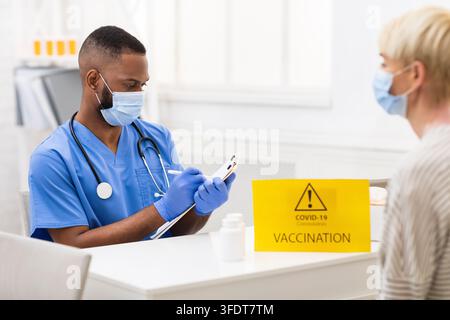 Covid-19 Vaccination. Black Doctor Having Appointment With Patient Lady Vaccinating For Coronavirus, Working Taking Notes Sitting In Hospital. Corona Stock Photo