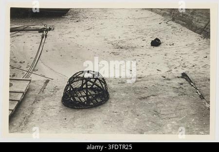 A black and white photograph by Geneviève Massignon, a French ethnologist, showing a domed, woven basket-like object, possibly related to aquaculture or fishing, on a sandy or gravelly ground, with a wooden structure and a metal frame in the background, taken between 1959 and 1965. Stock Photo