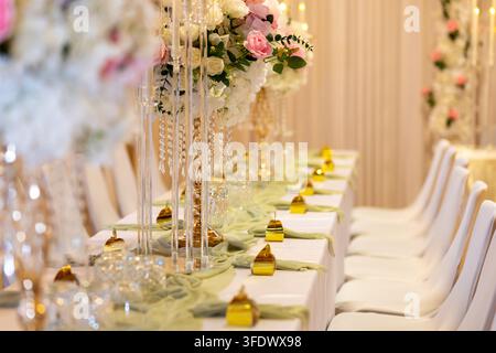 Long tables are set up in a bright room for a special event. Each table features floral arrangements and golden decorations with light shining on the Stock Photo