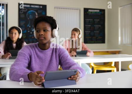 Diverse female youths sitting in classroom lavender sweater youth holding tablet wearing headphones Stock Photo