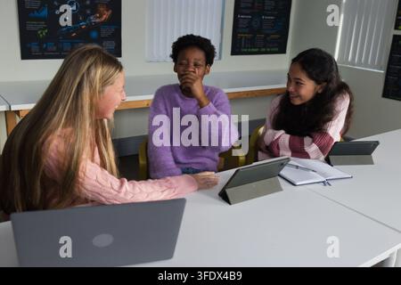 Female students laughing and studying together at classroom table, using tablets laptop notebook Stock Photo