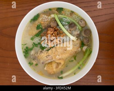 A white bowl of clear broth with chicken pieces, meatballs, rice, green onions, herbs, and crispy fried garnish sits on a wooden table. Stock Photo
