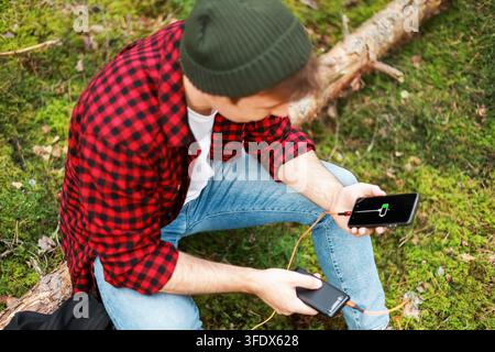 man resolves a battery emergency and power shortage outdoors in a forest, charging his smartphone with a portable power bank to stay connected. High q Stock Photo