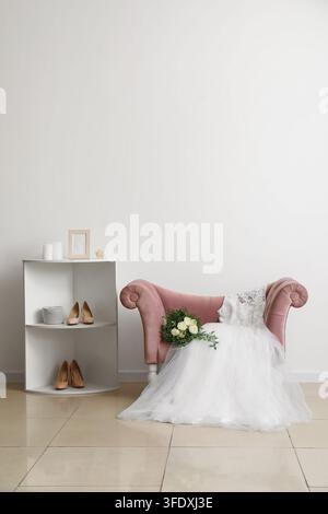 Wedding dress, bridal bouquet on pink armchair and wedding shoes on shelf near white wall in room Stock Photo
