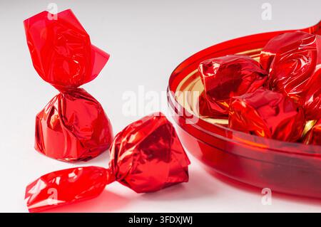 Red foil wrapped chocolate candies on a white surface next to a red bowl Stock Photo