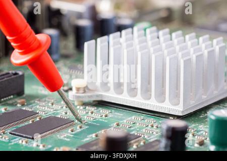 Red multimeter probe testing electronic components on a green circuit board with a silver heatsink Stock Photo