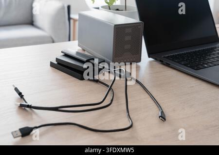 Stack of portable external hard drives and a two bay RAID enclosure connected by USB cables to a laptop on a wooden desk. Data backup and redundant Stock Photo
