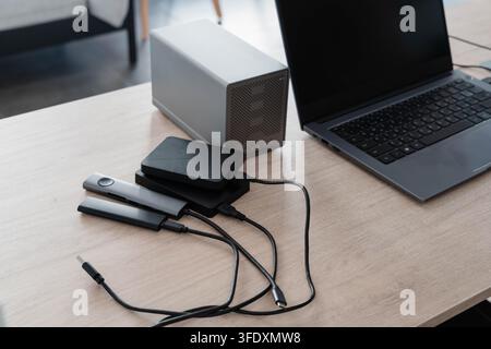 External drives, a network storage enclosure and a USB hub connect by cable to an open laptop on a wooden desk. Data backup and cloud storage sync in Stock Photo