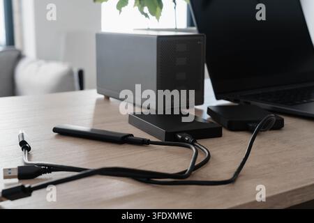 External hard drives, a portable SSD and USB cables connect to a laptop on a wooden desk. Home office setup for data backup, file archiving and secure Stock Photo
