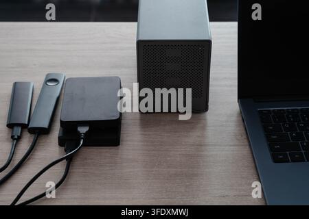 Portable external hard drives and SSD enclosures connected by cables to a laptop on a wooden desk. Data storage and backup workflow for photographers Stock Photo