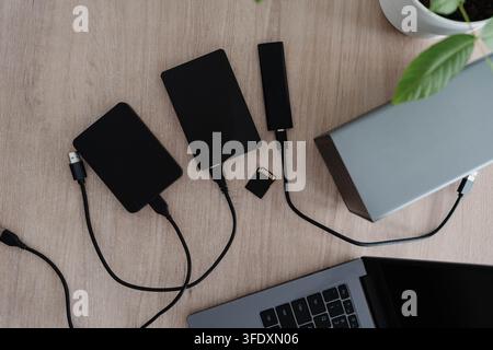 External hard drives, a portable SSD and a memory card lie with cables on a wooden desk beside an open laptop. Data storage and backup setup for Stock Photo