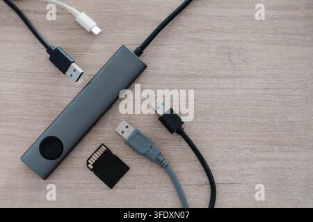 Aluminum USB hub with a glowing indicator lies on a wooden desk beside connector cables and a memory card. Peripheral connectivity and data transfer Stock Photo