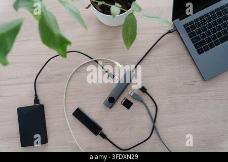 USB hub connected to a laptop on a wooden desk with an external hard drive, portable SSD, memory card and unplugged cables around it. Expanding ports Stock Photo