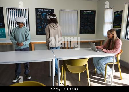 Diverse female teenage students wearing VR headsets and using laptop in classroom with AI posters Stock Photo