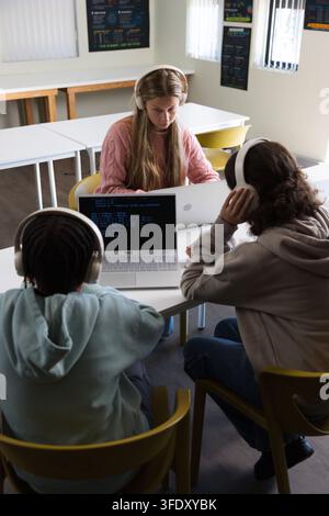 Youth female students sitting in small study room coding on laptops with headphones Stock Photo