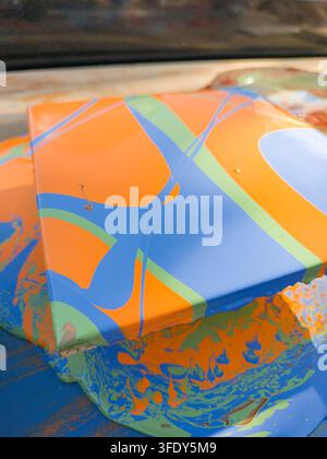 Vibrant paint splashes cover a glossy, geometric surface. Bold orange, blue, and lime swirls create dynamic visual energy. Colors bleed and blend, cap Stock Photo
