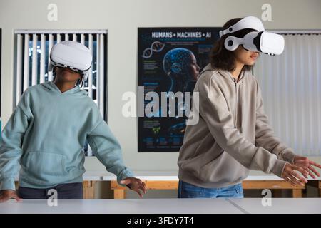 Diverse teenage females using white VR headsets in classroom lab with long table and AI poster Stock Photo