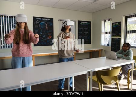 Diverse students wearing white VR headsets, gesturing at VR content at lab table with silver laptop Stock Photo