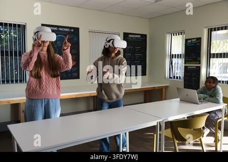 Diverse teenagers in pink sweater, gray hoodie gesturing with VR headsets, peer using laptop in lab Stock Photo