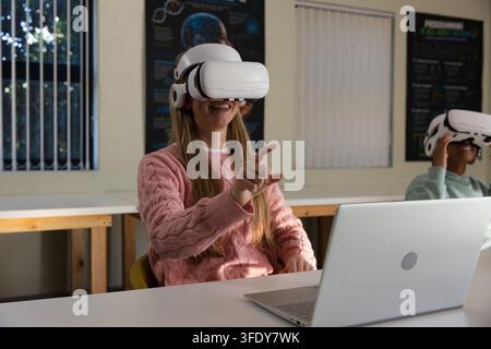 Diverse teenage students female in pink knit using white VR headsets interacting with laptop in lab Stock Photo