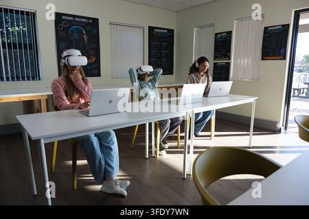 Diverse students sitting at white table in school lab using silver laptops, VR headsets, headphones Stock Photo