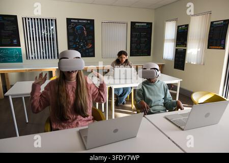 Youth students sitting at tables in classroom wearing white VR headsets using laptops Stock Photo