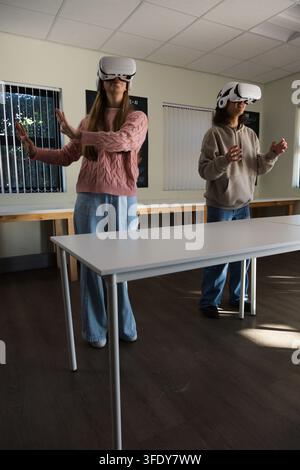 Diverse female youths wearing white VR headsets, standing behind white table in classroom gesturing Stock Photo