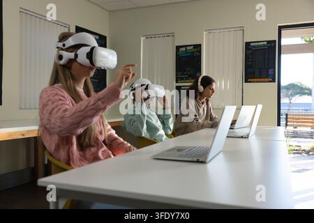 Diverse teen classmates sitting at white table in computer lab using VR headsets, laptops Stock Photo