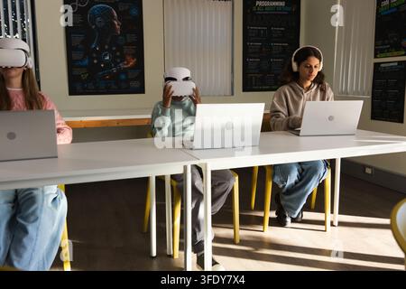 Diverse youth students wearing VR headsets, adjusting headset, typing on laptops in classroom Stock Photo