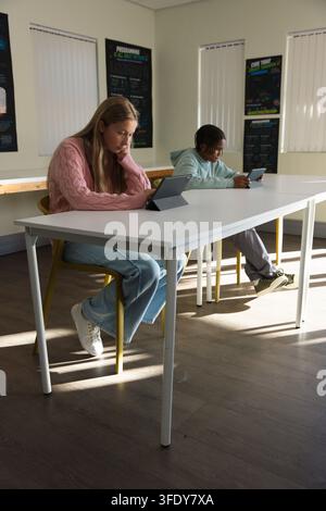 Diverse teen classmates sitting at long white table holding tablets on folio case Stock Photo