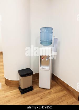 White water cooler with a plastic bottle and cup holder positioned in an office corner beside a small waste bin on a wooden floor, hydration station, indoor utility Stock Photo