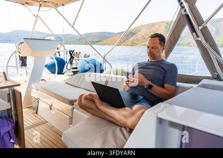 Man working remotely on a sailing yacht with laptop and smartphone. Location independent business and flexible lifestyle concept Stock Photo