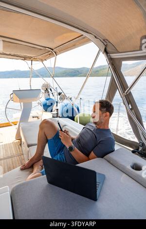 Man working remotely on a sailing yacht with laptop and smartphone. Location independent business and flexible lifestyle concept Stock Photo
