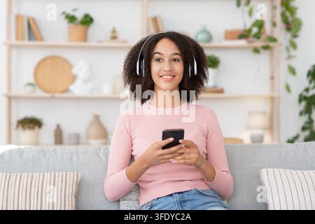 Mixed race woman in headphones holds smartphone, resting on couch Stock Photo