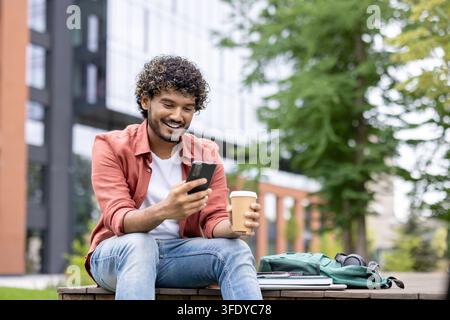 Smiling young man with curly hair sitting outdoors on a university campus, holding a coffee cup and engrossed in his smartphone, with a backpack and books nearby Stock Photo
