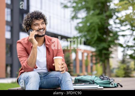 Young man on a university campus bench talking on his smartphone while holding a takeaway coffee, smiling and enjoying a relaxed outdoor break between classes or remote work sessions Stock Photo