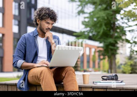 Young man using a laptop, contemplating ideas and focusing on work or study outside a modern building, representing remote work, flexible learning, and urban freelance lifestyle Stock Photo