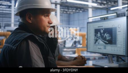 In Glass Control Room, Two Engineers Supervise Automated Robotic Assembly Lines on Dual Displays. Cyber Physical Systems and Smart Automation Drive High Tech Plant Efficiency. Team Collaboration. Stock Photo